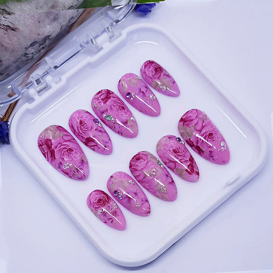 A set of pink nails with a floral design and some 3d elements, displayed in a white case.
