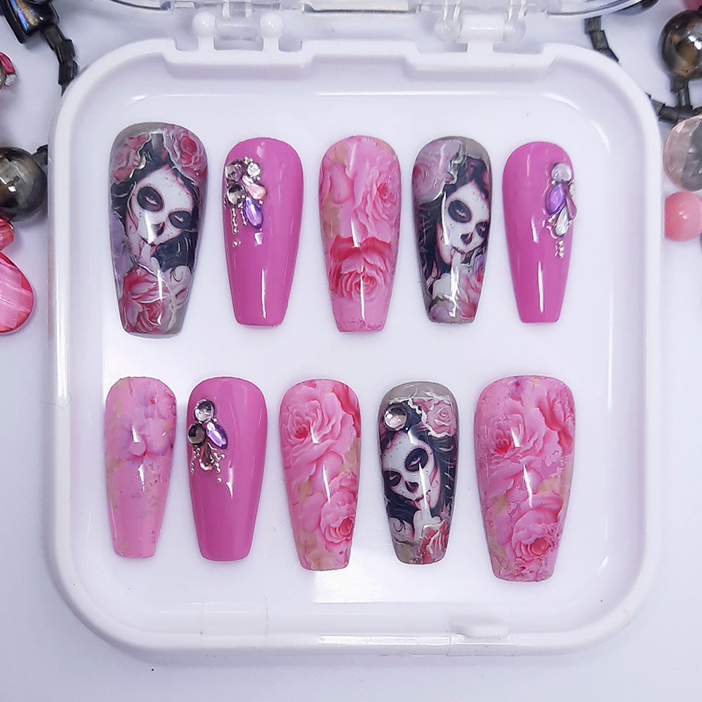 A set of pink and grey press on nails with a floral and faces design and some 3d nail art, displayed in a case on a white background.
