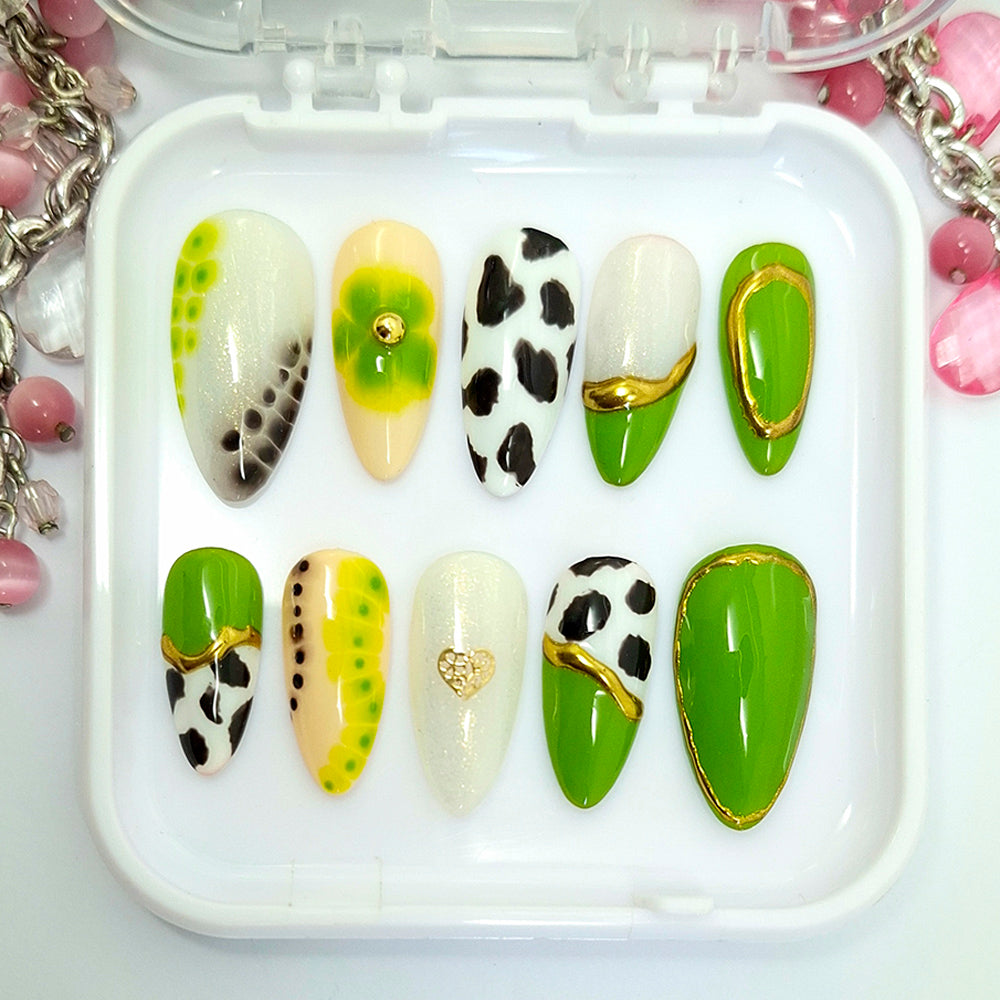 A set of ten press-on nails displayed in a clear plastic tray. The nails feature various designs including green and white gradients, black and white cow print, gold accents, and splatter patterns.