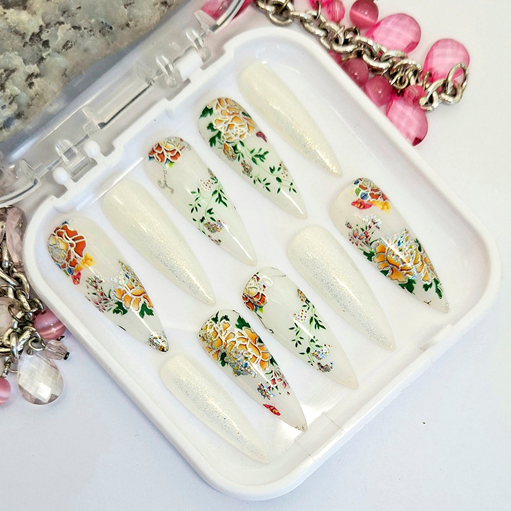 A set of milky white and glitter long stiletto press on nails with an oriental design of colourful flowers and leaves, displayed in a white case on a white background.