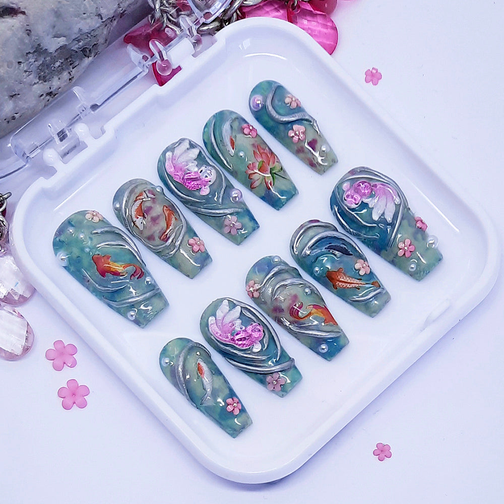 A set of press on nails in blue/green with an elaborate design of koi fish and flowers including some 3d nail art, displayed in a white tray on a white background.