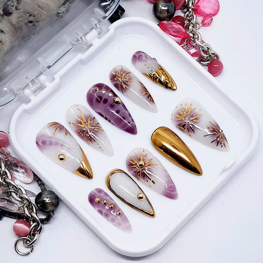 Set of decorative nail art tips with gold and purple designs in a white tray.