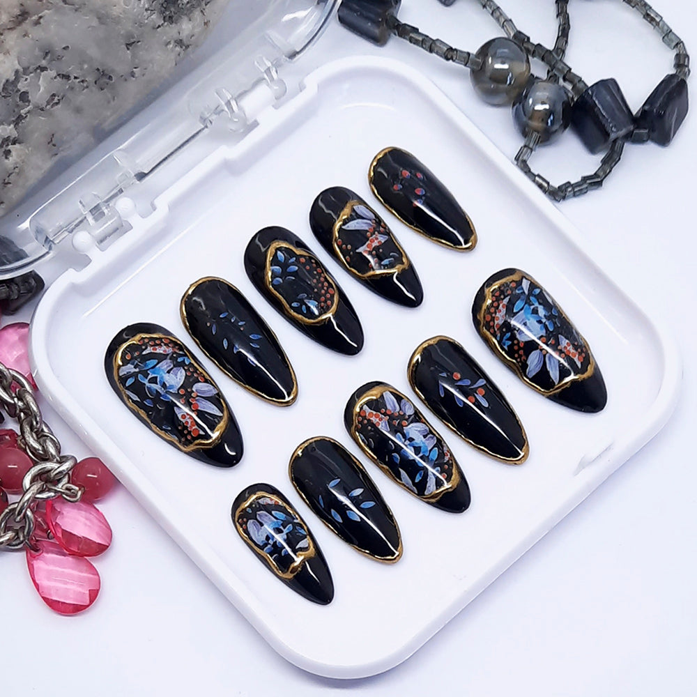 A set of black press on nails with patches of abstract designs of leaves and berries with gold chrome displayed in a white tray on a white background.
