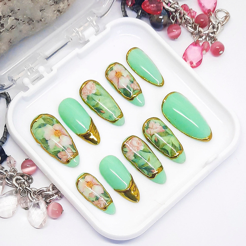 A set of green press on nails with a floral design and gold chrome, displayed in a white tray on a white background.