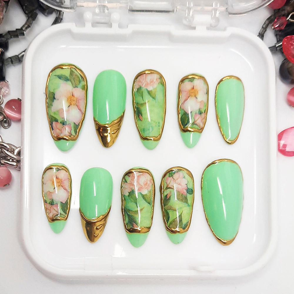 A set of green press on nails with a floral design and gold chrome, displayed in a white tray on a white background.