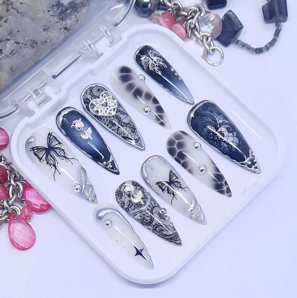 A set of black and white long press on nails, with multiple designs including lace, butterflies and silver 3d nail art. Displayed in a white tray on a white background.