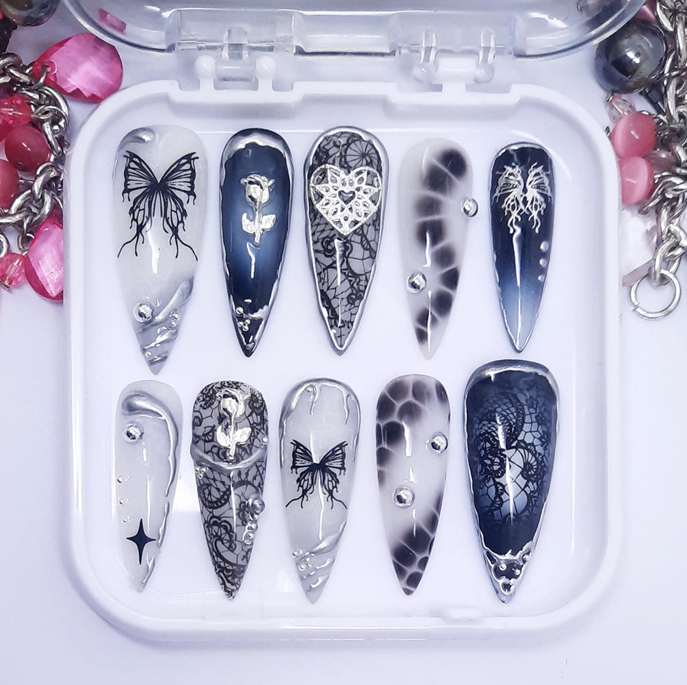 A set of black and white long press on nails, with multiple designs including lace, butterflies and silver 3d nail art. Displayed in a white tray on a white background.