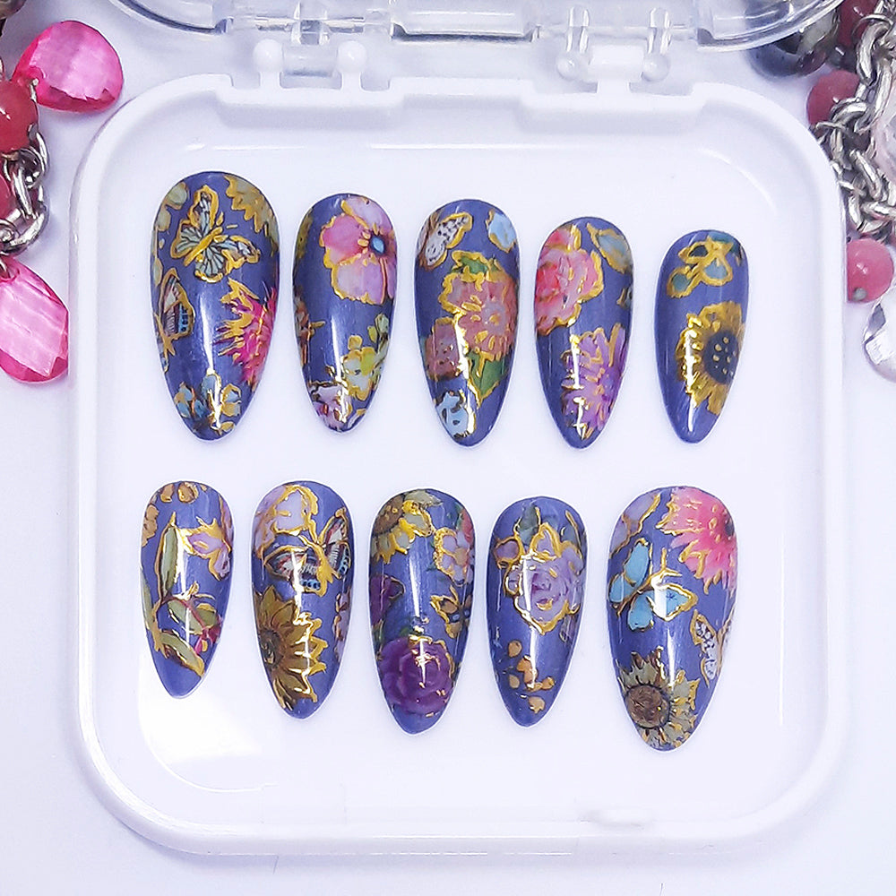 A set of press on nails with a colourful design of flowers and butterflies with gold detail, displayed in a white tray on a white background.
