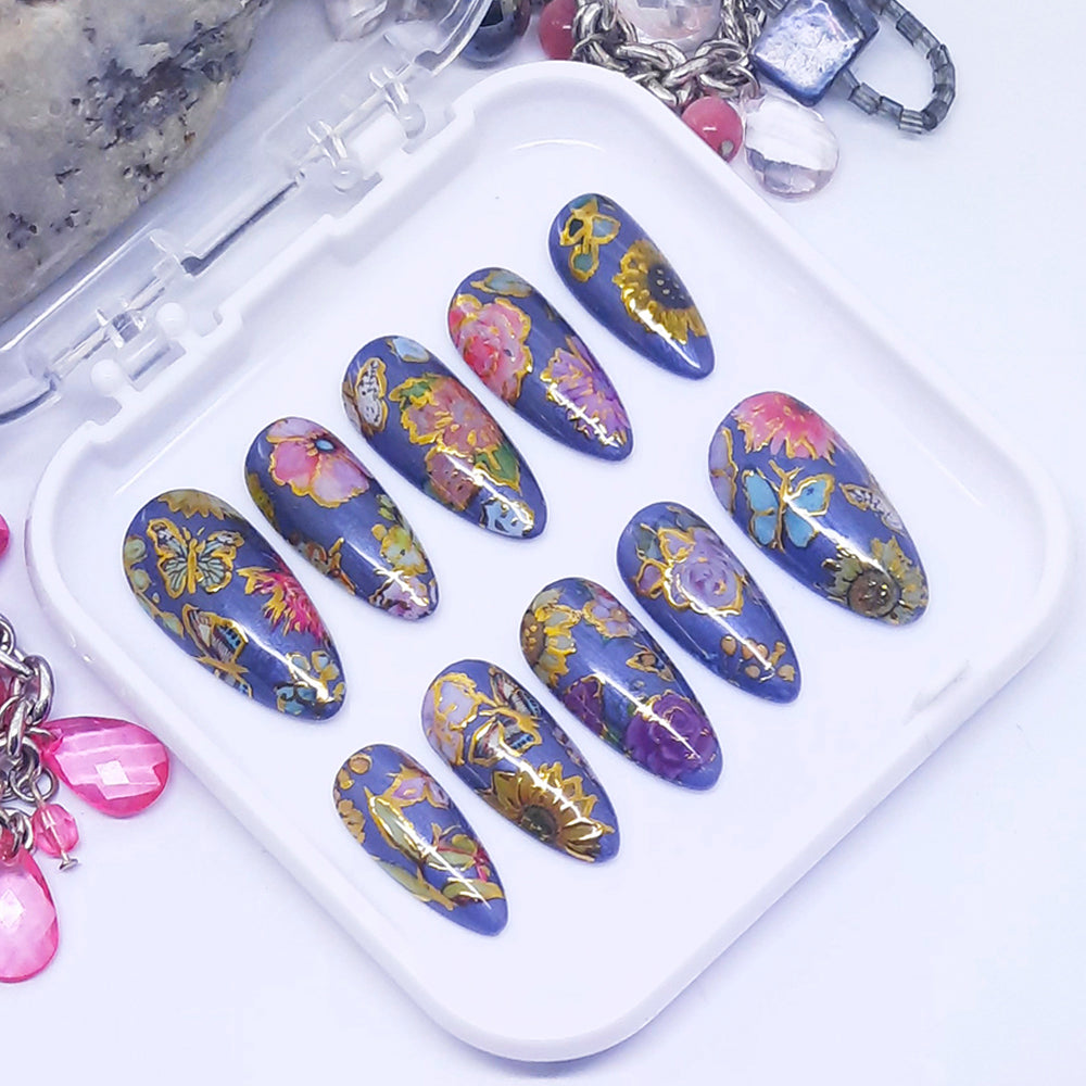 A set of press on nails with a colourful design of flowers and butterflies with gold detail, displayed in a white tray on a white background.