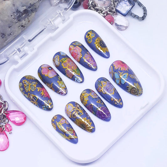 A set of press on nails with a colourful design of flowers and butterflies with gold detail, displayed in a white tray on a white background.