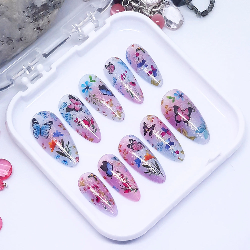 A set of pink and blue themed press on nails with a floral and butterfly design displayed in a white tray n a white background.