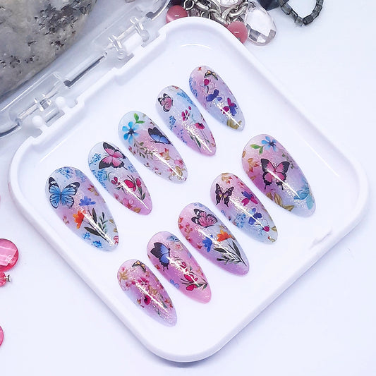 A set of pink and blue themed press on nails with a floral and butterfly design displayed in a white tray n a white background.