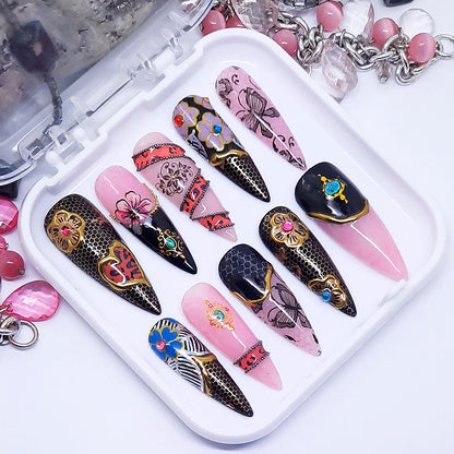 A set of long stiletto nails in a pink and black theme with multiple nail decoration including flowers, butterflies and gold chrome. Displayed in a white tray on a white background.