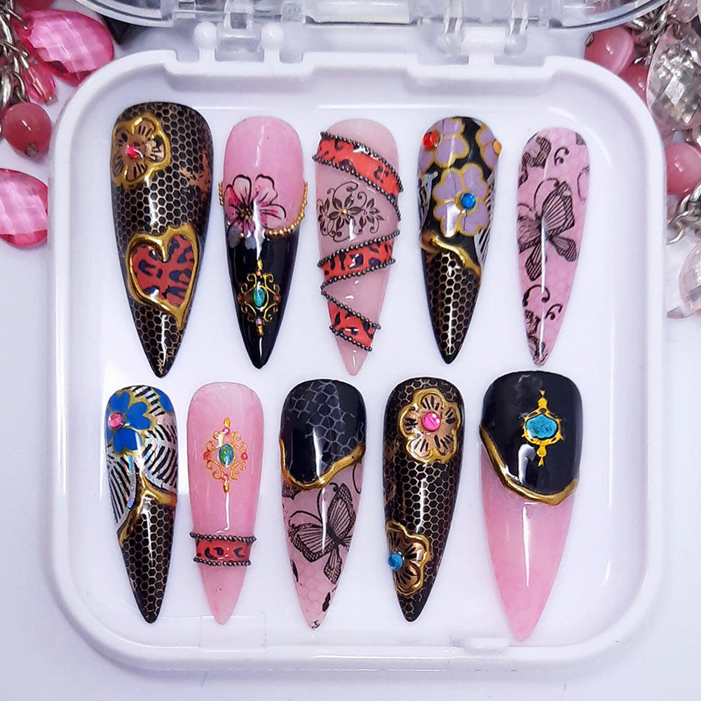 A set of long stiletto nails in a pink and black theme with multiple nail decoration including flowers, butterflies and gold chrome. Displayed in a white tray on a white background.