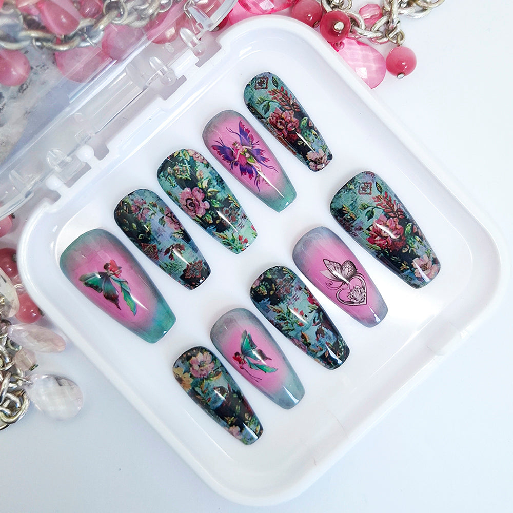 A set of hand made press on nails in pink, blue and green shades with a design of flowers, fairies and a butterfly. Displayed in a white tray on a white background.