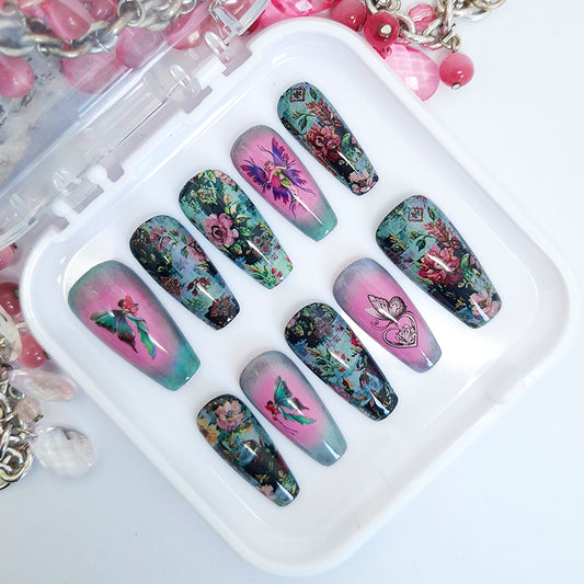 A set of hand made press on nails in pink, blue and green shades with a design of flowers, fairies and a butterfly. Displayed in a white tray on a white background.