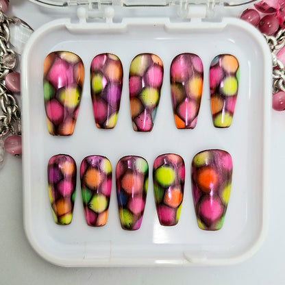A set of very colourful medium coffin press on nails with a design of multi-coloured patches and finished with a chrome sheen. Displayed in a white tray on a white background.