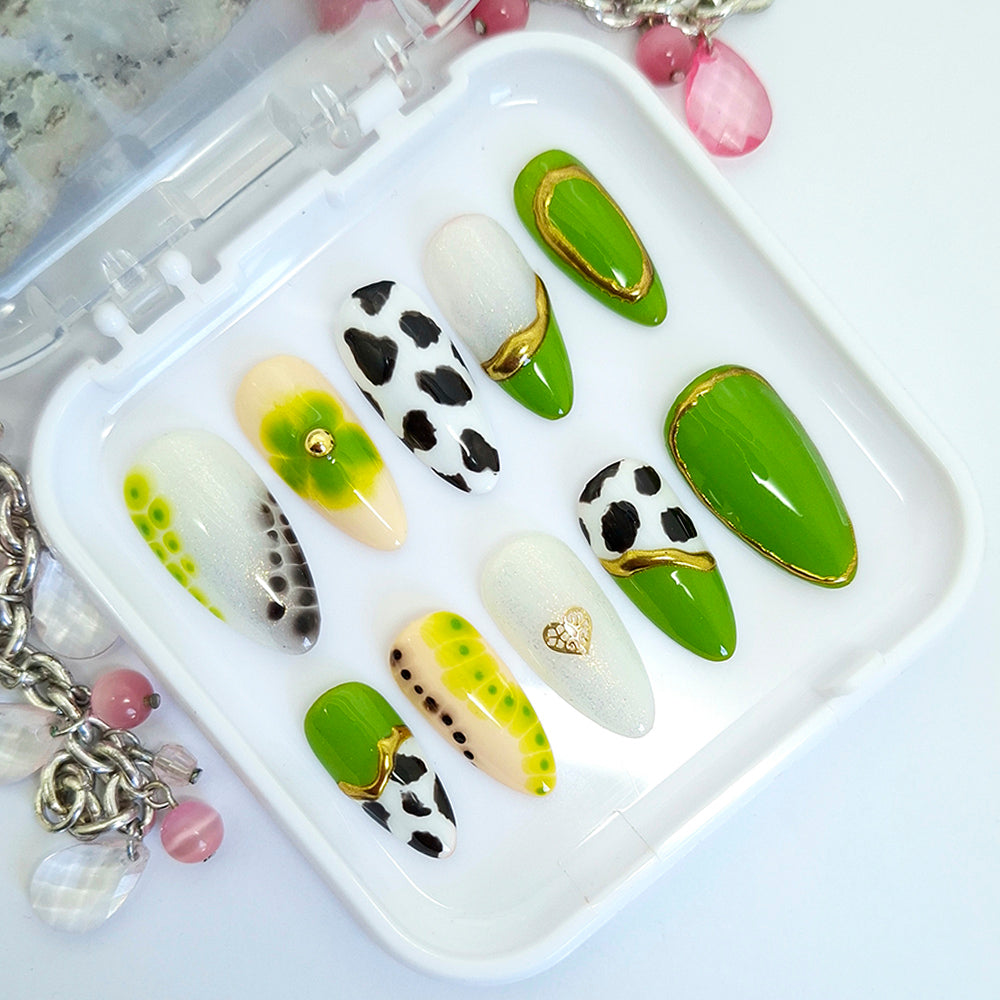 A set of 10 press-on false nails displayed in a clear plastic tray. The nails feature various designs including solid green, white with black cow print, green and yellow gradient, and gold heart and wave details.