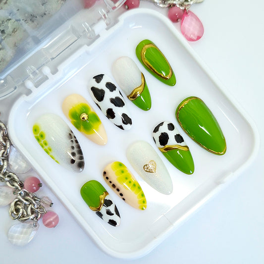 A set of 10 press-on false nails displayed in a clear plastic tray. The nails feature various designs including solid green, white with black cow print, green and yellow gradient, and gold heart and wave details.