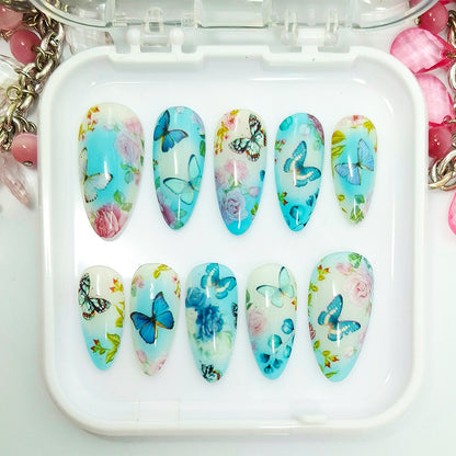 A set of 10 artificial press-on nails displayed in a clear plastic case, featuring a light blue and white background decorated with blue butterflies, pink roses, and green leaves.