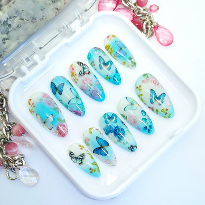 A set of artificial press-on nails with butterfly and floral designs, displayed in a white plastic case on a white surface with decorative jewelry props.
