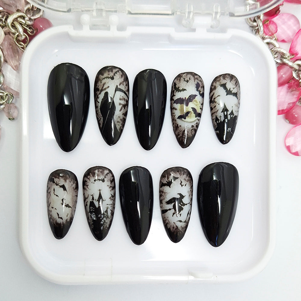 A set of ten almond-shaped press-on nails displayed in a clear plastic case on a white surface. The nails feature a black and white gothic Halloween design, including splatters, and bats with some nails having a glossy black finish and others showing a distressed white-and-black pattern.