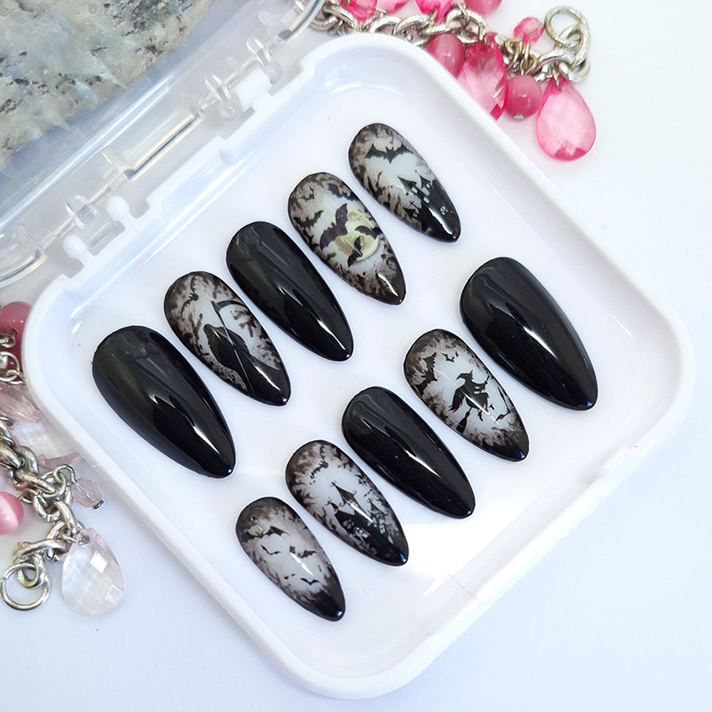 A set of ten almond-shaped press-on nails displayed in a clear plastic case on a white surface. The nails feature a black and white gothic Halloween design, including splatters, and bats with some nails having a glossy black finish and others showing a distressed white-and-black pattern.