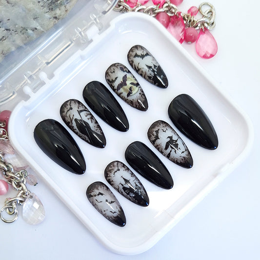 A set of ten almond-shaped press-on nails displayed in a clear plastic case on a white surface. The nails feature a black and white gothic Halloween design, including splatters, and bats with some nails having a glossy black finish and others showing a distressed white-and-black pattern.