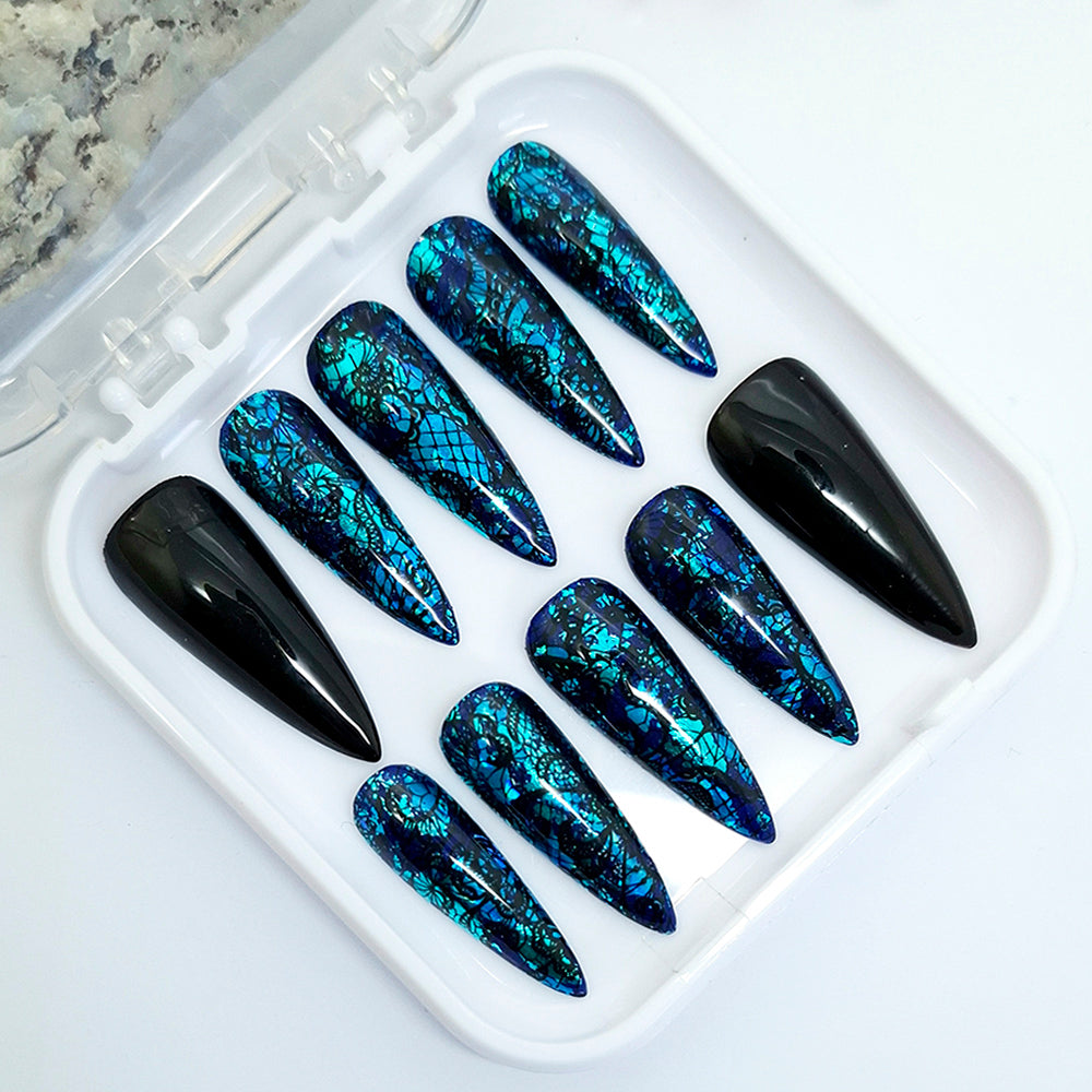 A set of ten stiletto press-on nails displayed in a white plastic tray. Nine nails feature a glossy black base with shimmering blue and teal metallic crackle patterns, while one nail is solid glossy black.